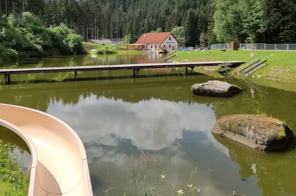 Holzsteg quer über den Waldbadteich, dahinter das Waldbadgebäude