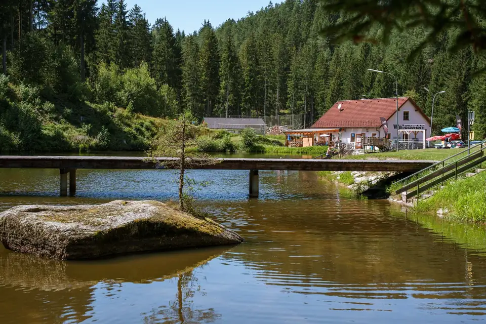 Waldbadteich mit Holzsteg, Granitfelsen, Hauptgebäude und dichtem Wald im Sommer